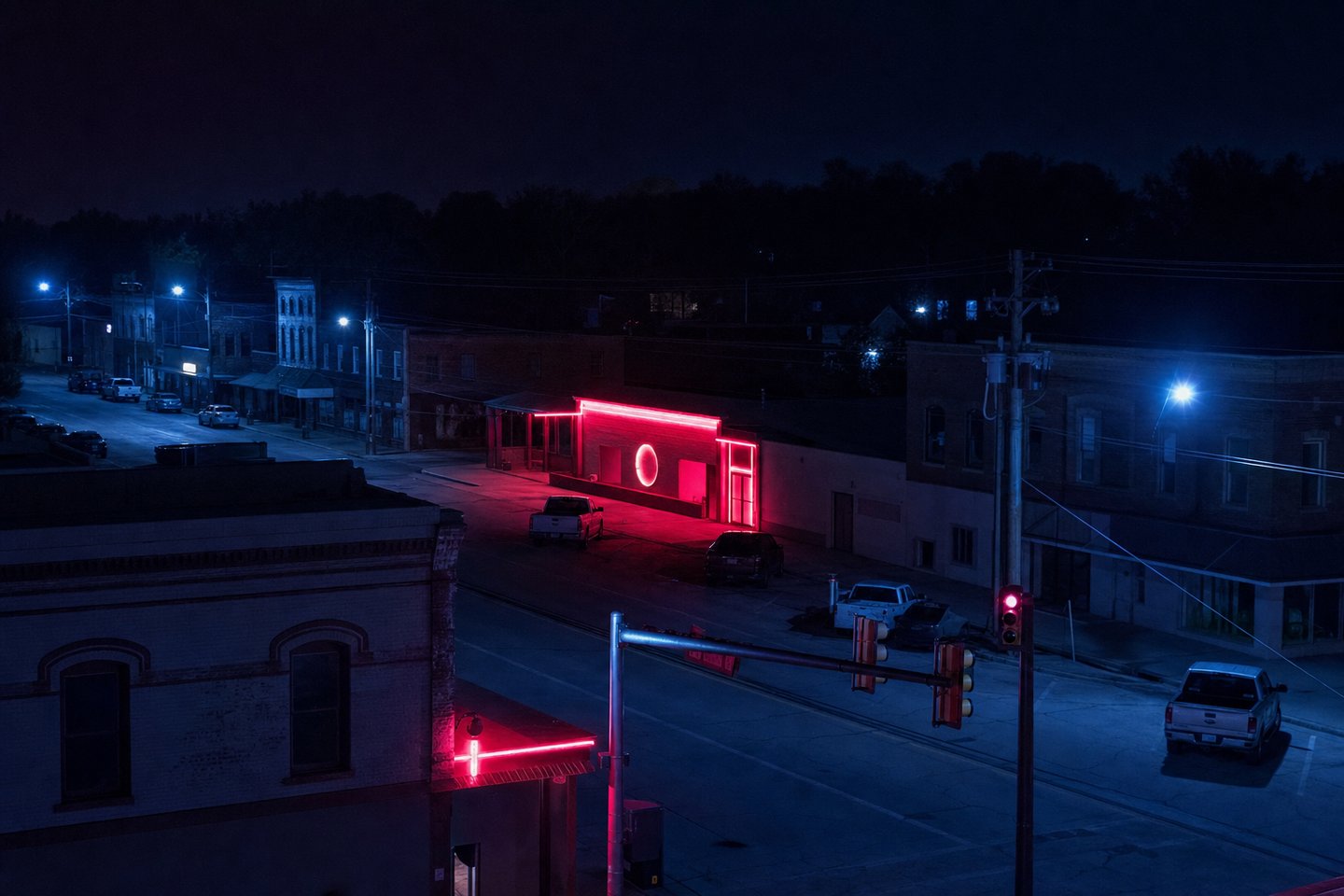 An elevated night view of an ordinary American small-town main street, most storefronts dark, with a single-storey casino among them lit in red neon and pickup trucks parked along the kerb.