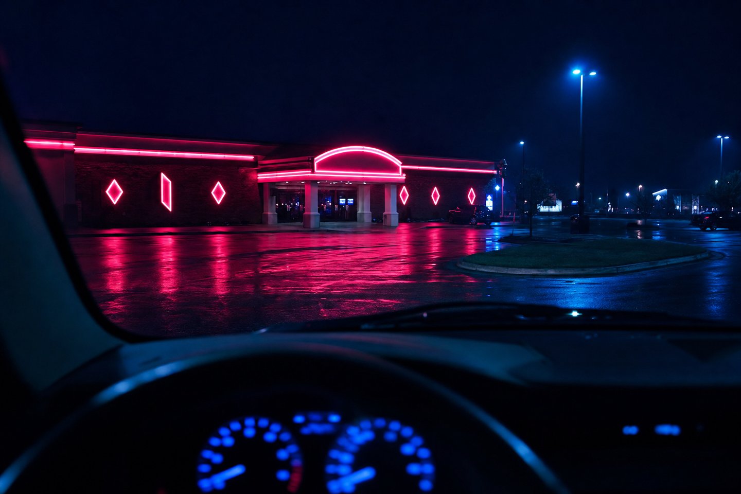 The view from a car's driver seat at night: dashboard instruments glowing blue in the foreground and, through the windscreen, a low casino building lit in red neon across a wet car park.